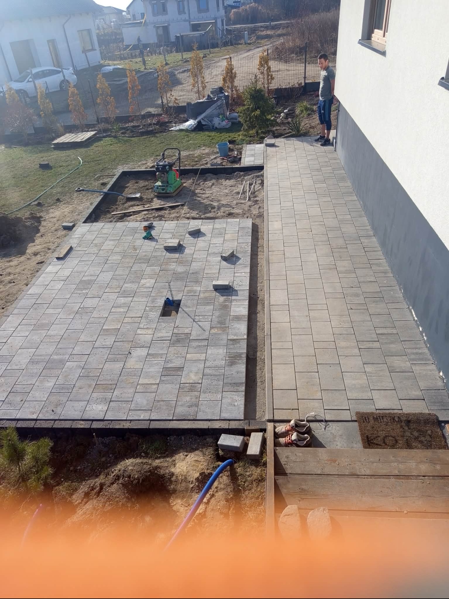 Fragment układanej szarej kostki brukowej na posesji, widoczny pracownik, zagęszczarka i przygotowany teren pod dalsze prace. Ujęcie z góry.