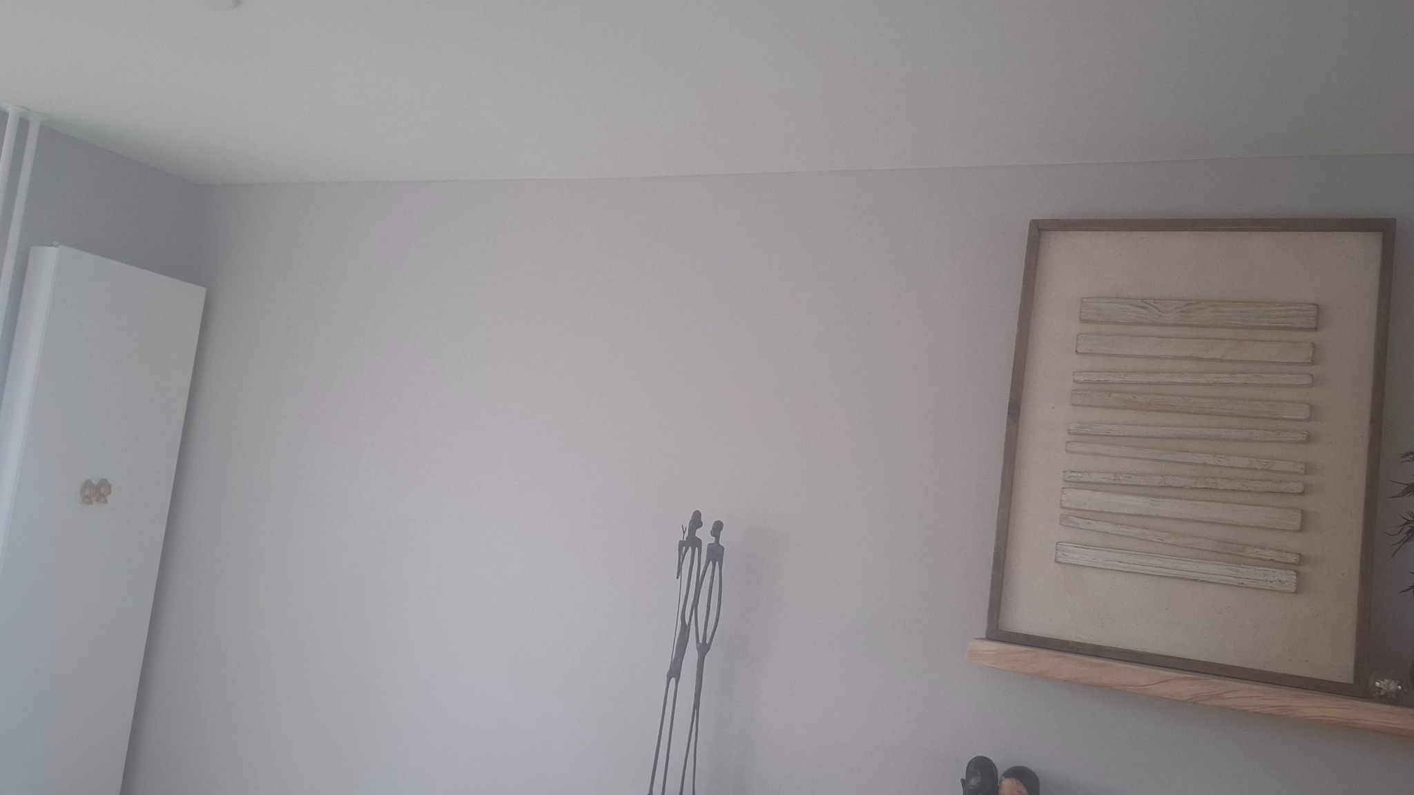 Jasnoszara ściana po malowaniu z dekoracjami: obraz w ramie i metalowa figurka. Widoczny fragment białej szafy z ozdobnymi uchwytami.