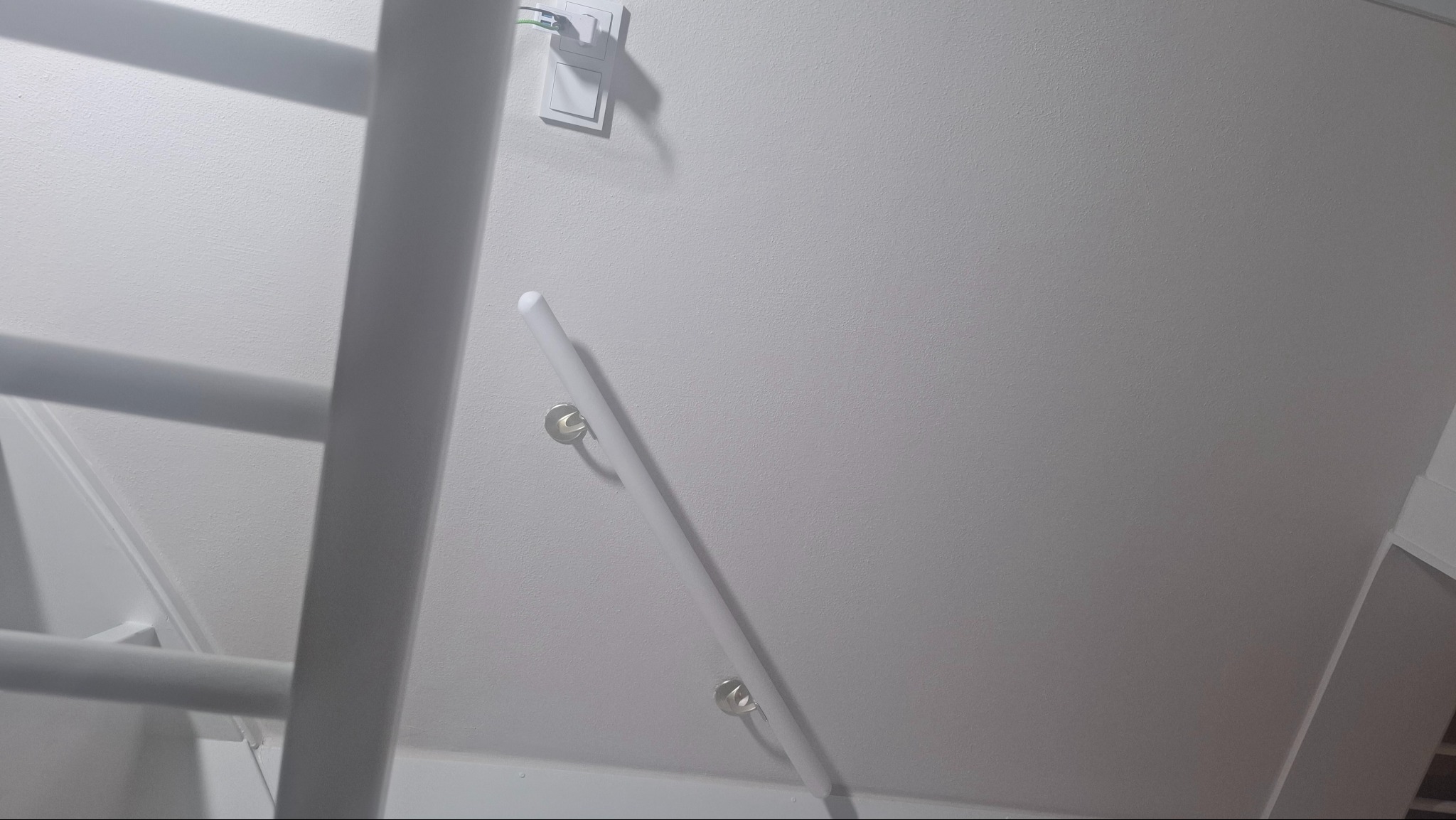 Biała, świeżo pomalowana ściana z zamontowaną poręczą i gniazdkiem elektrycznym. Fragment drabiny malarskiej w lewym rogu kadru. Tekstura ściany widoczna.
