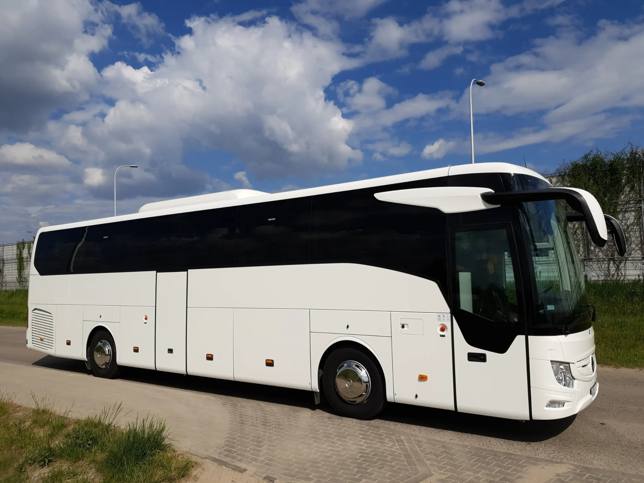 Nowoczesny autokar turystyczny Mercedes w kolorze białym, zaparkowany na tle błękitnego nieba z chmurami. Widok z boku, oświetlenie dzienne.
