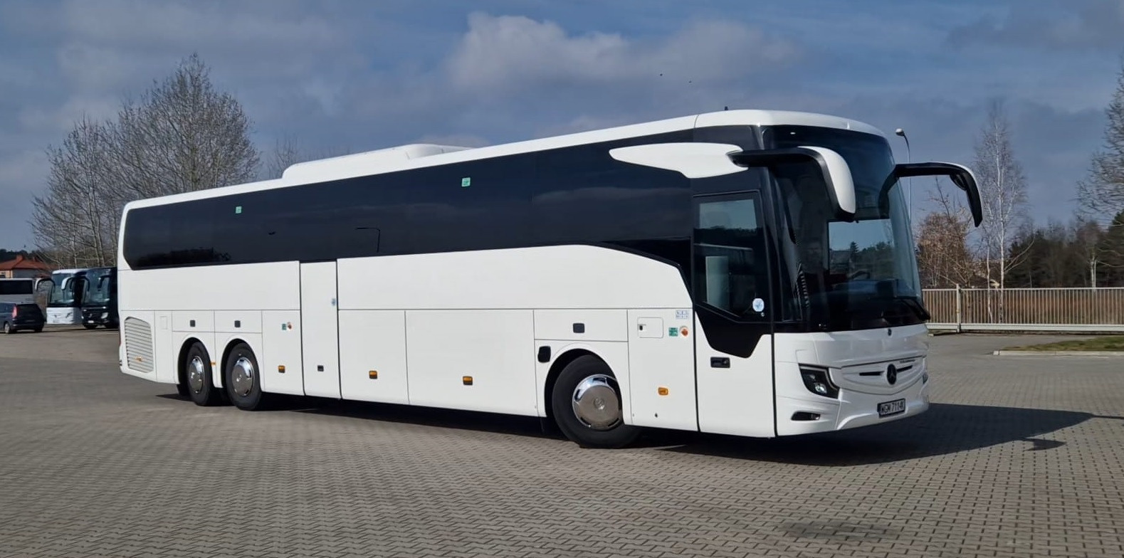 Nowoczesny, biały autokar Mercedes-Benz z czarnymi szybami, zaparkowany na placu z kostki brukowej. Widoczne detale: chromowane felgi, otwierane lusterko boczne.