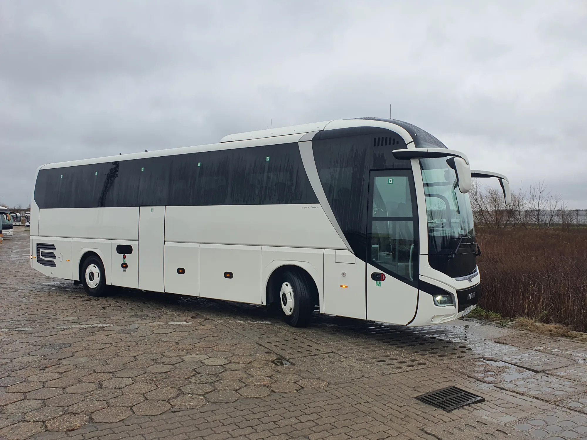 Nowoczesny, biało-czarny autokar turystyczny marki MAN na parkingu z kostki, widoczny z boku, pochmurne niebo.