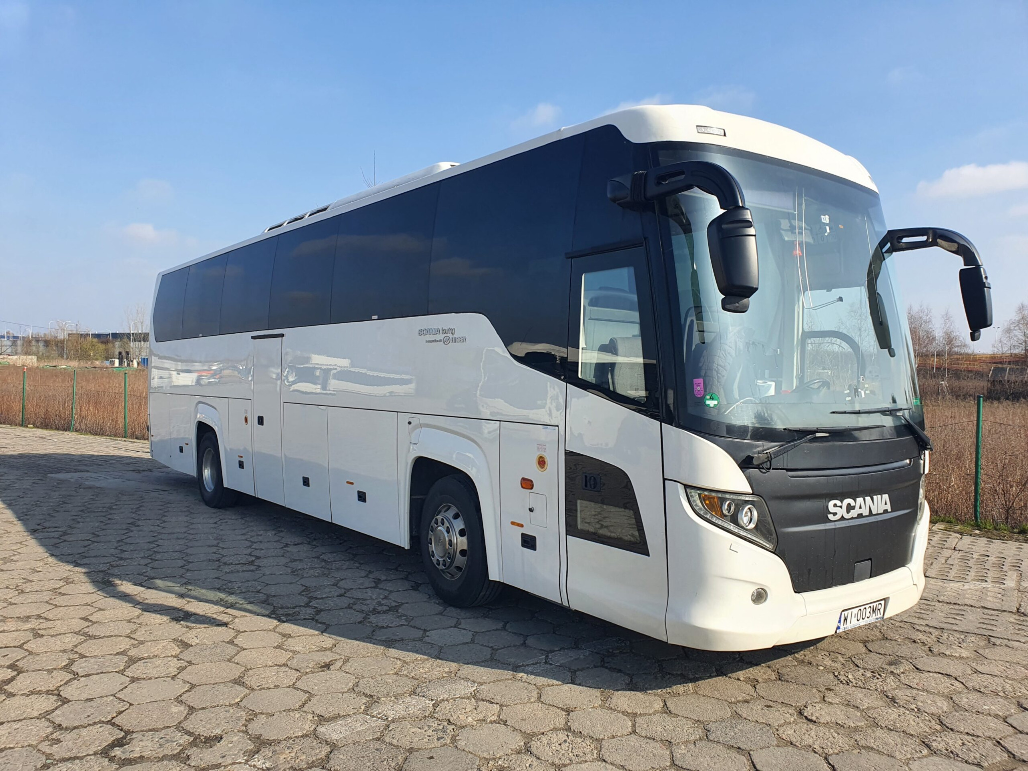 Nowoczesny, biały autokar Scania z czarnymi szybami i elementami, zaparkowany na kostce brukowej w słoneczny dzień. Widoczny numer rejestracyjny i logo producenta.