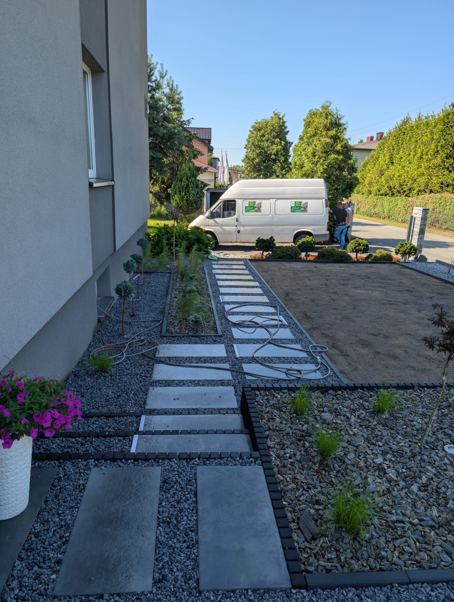 Nowoczesny ogród po założeniu: ścieżka z płyt, rabaty z kamykami i roślinami, trawnik przygotowany pod zasiew. Na podjeździe biały bus z logo firmy.