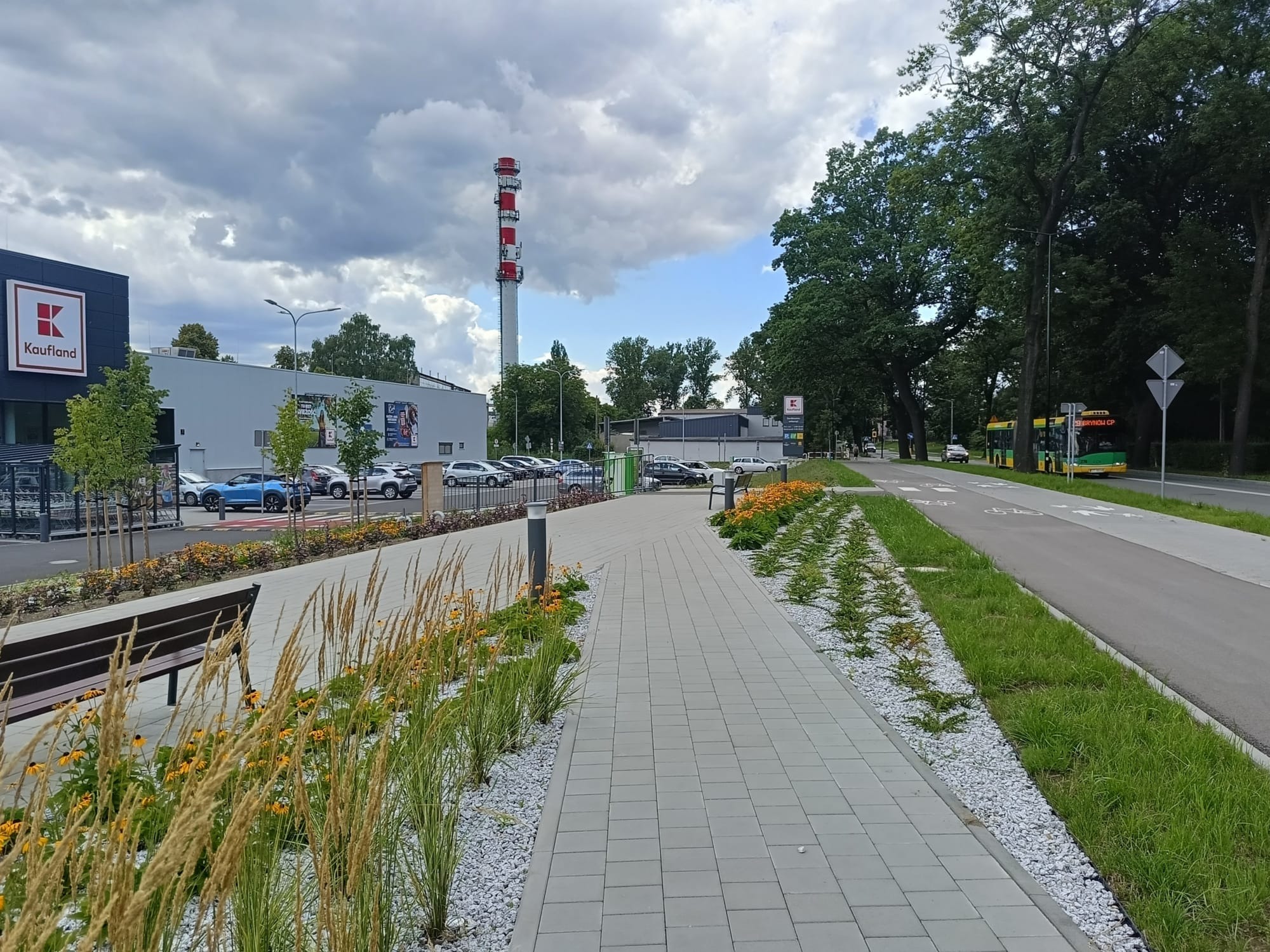 Ścieżka z kostki brukowej obok parkingu sklepu, z rabatami kwiatowymi i ozdobnymi trawami. W tle komin i budynek sklepu.