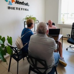 Paweł Nevada - Konsultacja dietetyczna: para siedzi przy biurku z dietetykiem, który prezentuje informacje na laptopie. W tle logo 'Nedzak, Dietetyka' i roślina doniczkowa.