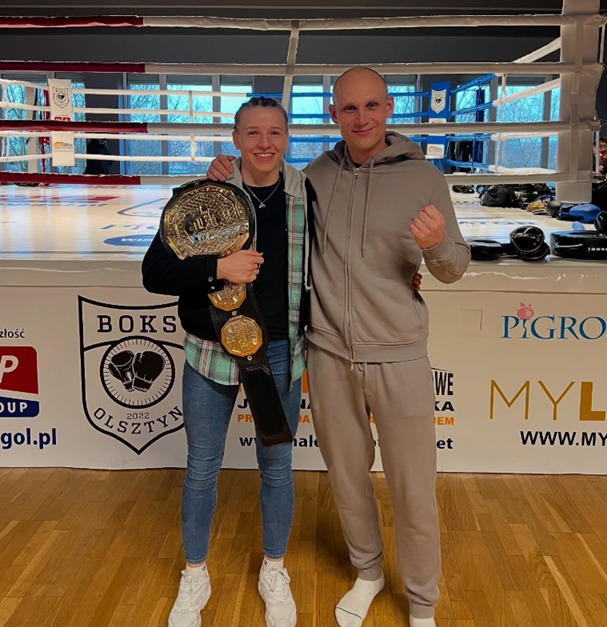 Uśmiechnięta kobieta z pasem mistrzowskim pozuje z trenerem na tle ringu bokserskiego. Widoczne logo klubu bokserskiego i sponsorów.