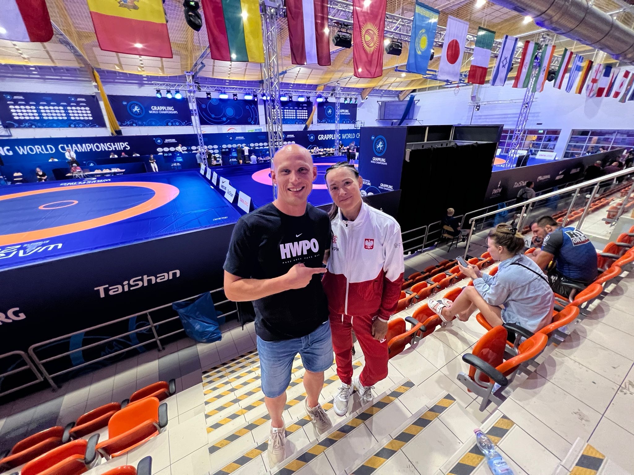 Dwoje sportowców na tle areny Grappling World Championships. Mężczyzna w koszulce HWPO wskazuje palcem. Kobieta w dresie reprezentacji Polski.