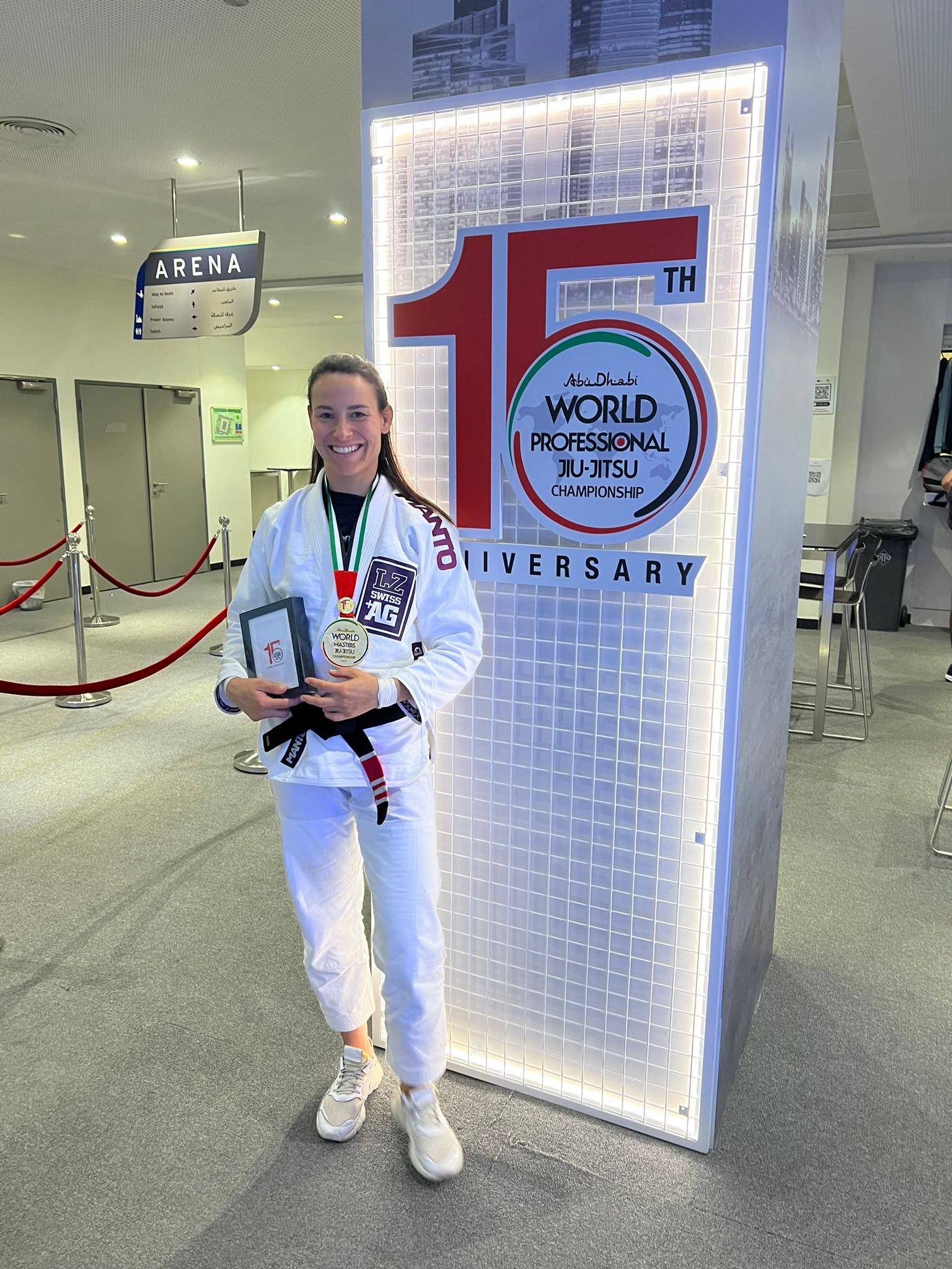 Uśmiechnięta kobieta w kimonie z czarnym pasem, z medalem i nagrodą na tle logotypu Abu Dhabi World Professional Jiu-Jitsu Championship.