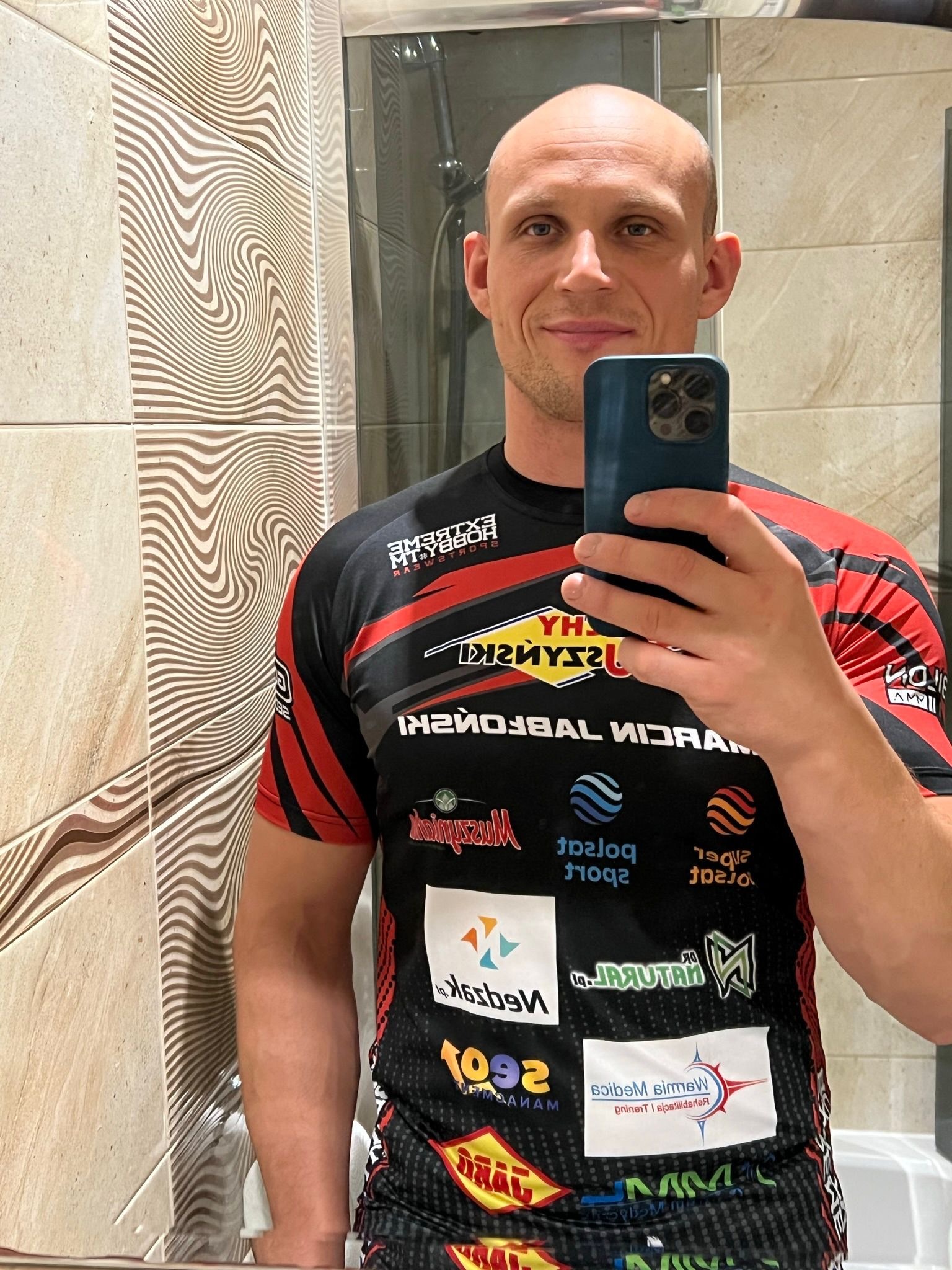 Mężczyzna robi selfie w lustrze łazienki, ubrany w sportową koszulkę z logotypami sponsorów. Widoczny uśmiech i telefon w dłoni.