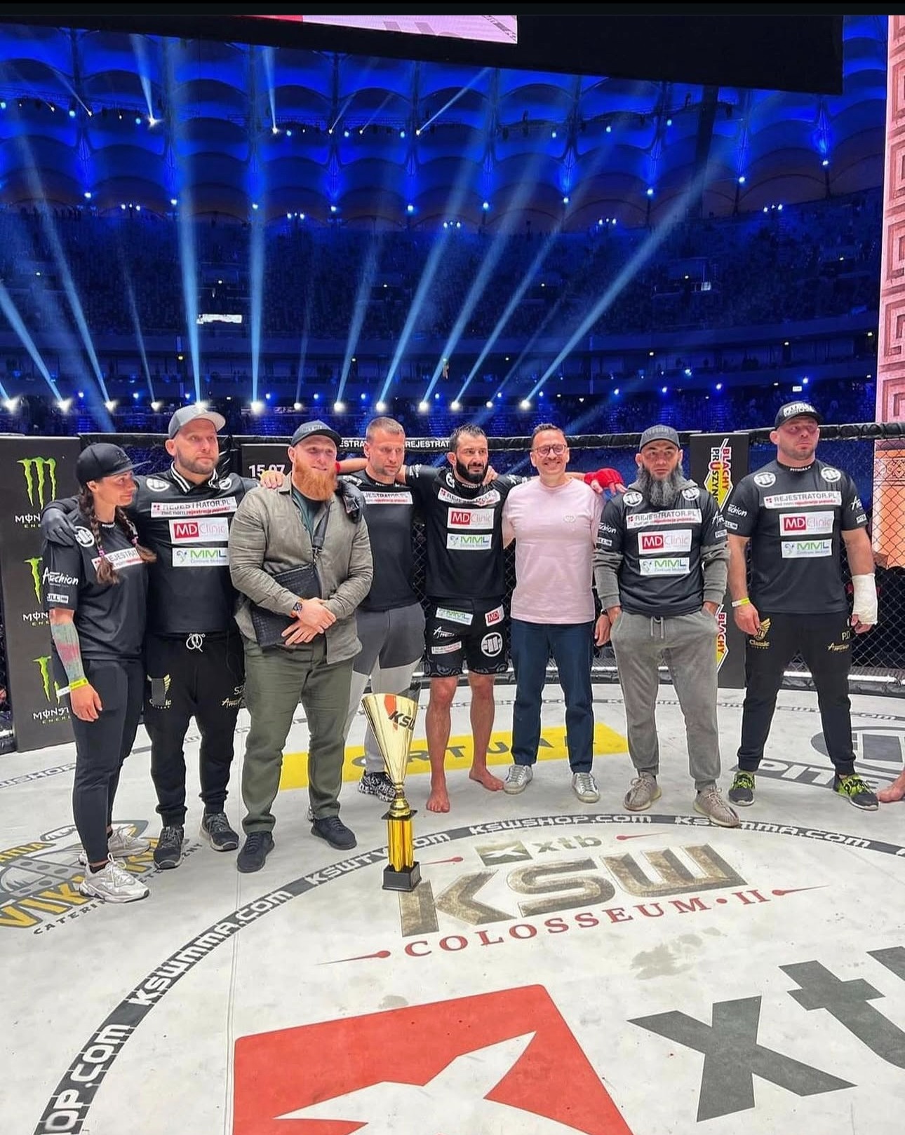 Grupa osób, w tym zawodnik MMA z trofeum, pozuje na ringu KSW. Widoczni sponsorzy i oświetlenie hali. Ujęcie z dołu, podkreślające atmosferę wydarzenia.