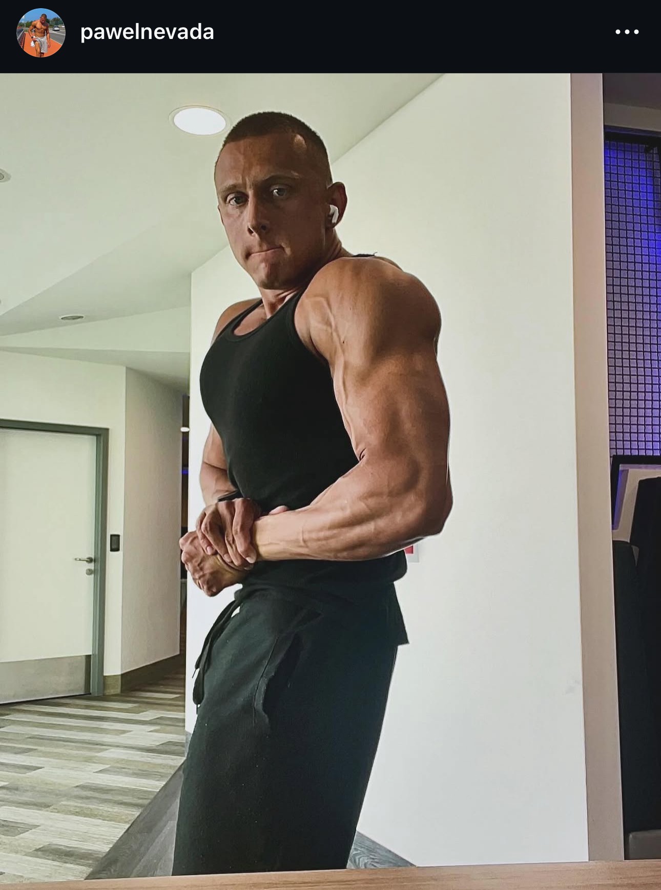 Mężczyzna w czarnym tank topie prezentuje muskulaturę ramion, stojąc na tle jasnej ściany i drzwi. Widoczne żyły na bicepsach, słuchawka w uchu. Ujęcie z dołu.