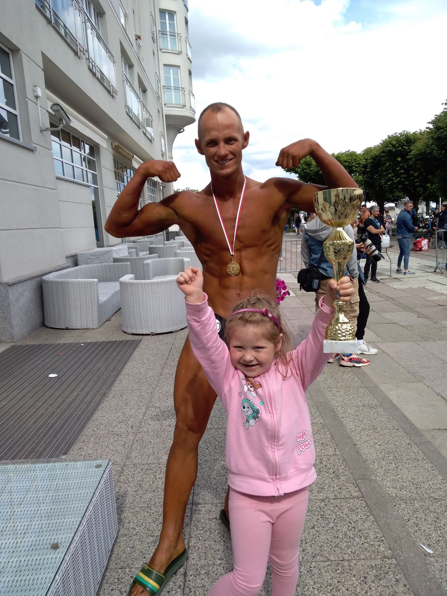 Uśmiechnięty kulturysta z medalem pozuje z dziewczynką trzymającą złoty puchar na tle miejskiej architektury i widzów. Widoczne detale: opalona skóra, sportowa sylwetka.