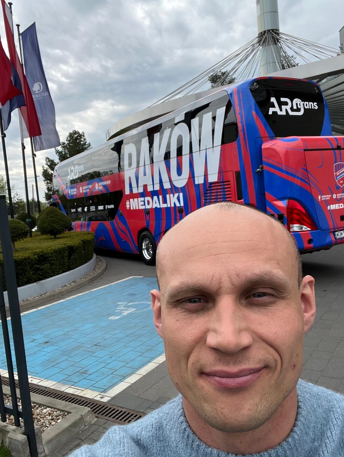 Selfie trenera na tle autobusu drużyny Raków Częstochowa. Widoczny budynek DoubleTree by Hilton i błękitne oznaczenia parkingu. Trener w niebieskim swetrze.