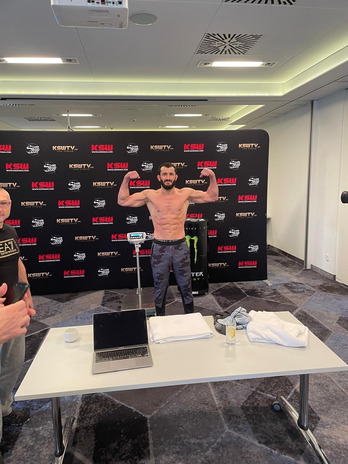 Mężczyzna prezentuje sylwetkę na wadze przed logotypem KSW. Widoczny laptop, ręczniki i woda z cytryną na stole. Przygotowania do ważenia zawodników MMA.