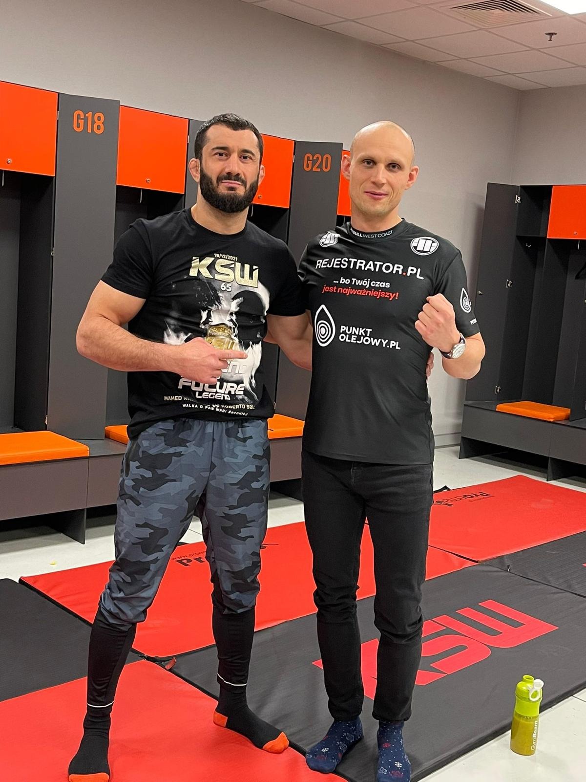 Dwóch mężczyzn w szatni, jeden w koszulce KSW, drugi w koszulce Rejestrator.pl, pozują do zdjęcia. W tle szafki z numerami G18 i G20.