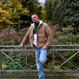 Mężczyzna w brązowej kurtce i okularach, oparty o metalową barierkę na tle jesiennej zieleni. Ujęcie portretowe, z wyraźnym tłem parkowym.