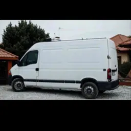 Biały bus dostawczy Renault Master na żwirowym podłożu, widok z boku, w tle zieleń i budynek z czerwoną dachówką. Lekko pochmurne niebo.