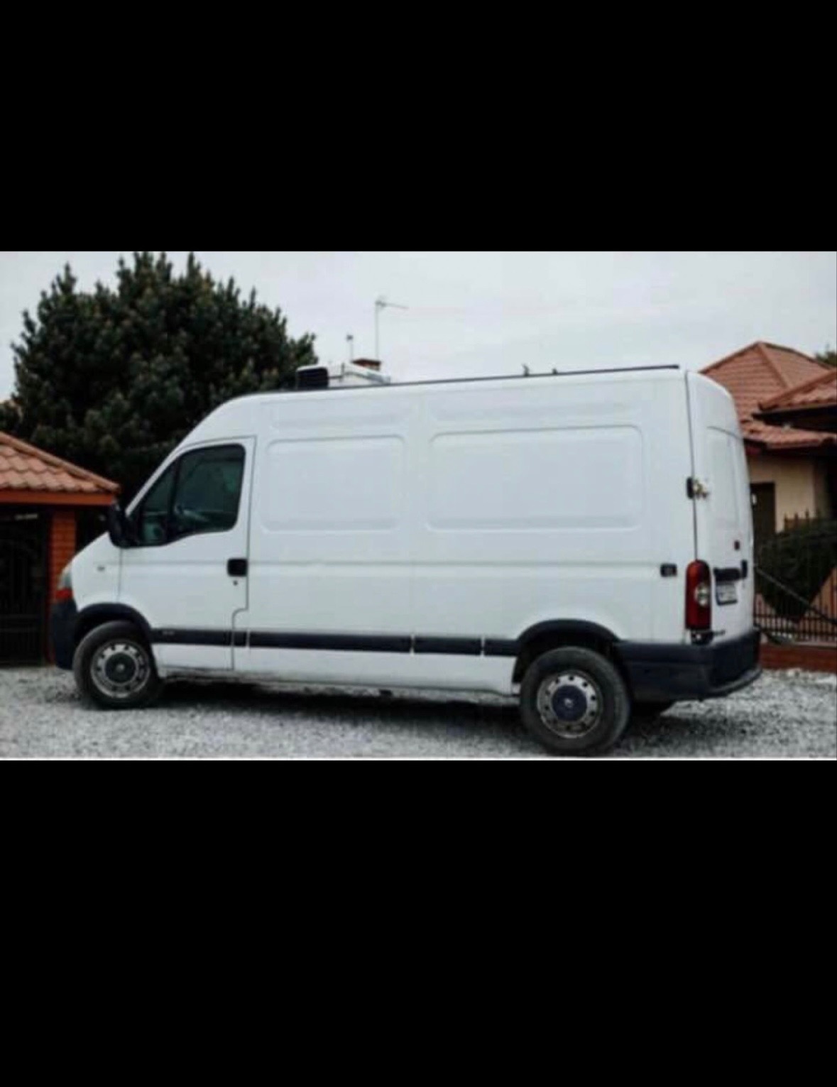 Biały bus dostawczy Renault Master na żwirowym podłożu, widok z boku, w tle zieleń i budynek z czerwoną dachówką. Lekko pochmurne niebo.