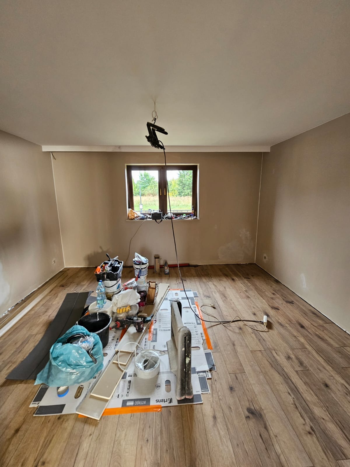 Pokój w trakcie remontu: świeżo ułożone panele podłogowe, pomalowane ściany na beżowo, narzędzia i materiały budowlane rozłożone na podłodze, widok na okno.