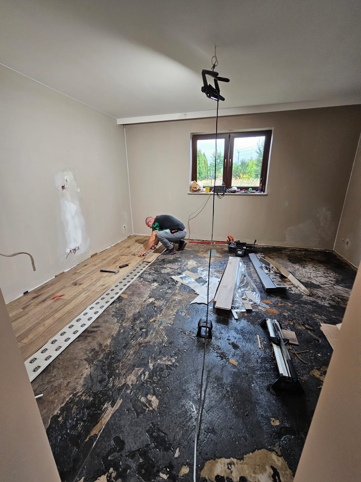 Mężczyzna w trakcie układania paneli podłogowych w pokoju. Widoczna część ułożonej podłogi, narzędzia i przygotowane panele. Ściany w jasnym odcieniu beżu.