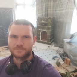 KoloMajstra - Mężczyzna z słuchawkami na szyi pozuje na tle remontowanego pomieszczenia z rozłożonymi kaflami i piecem kaflowym w trakcie budowy.