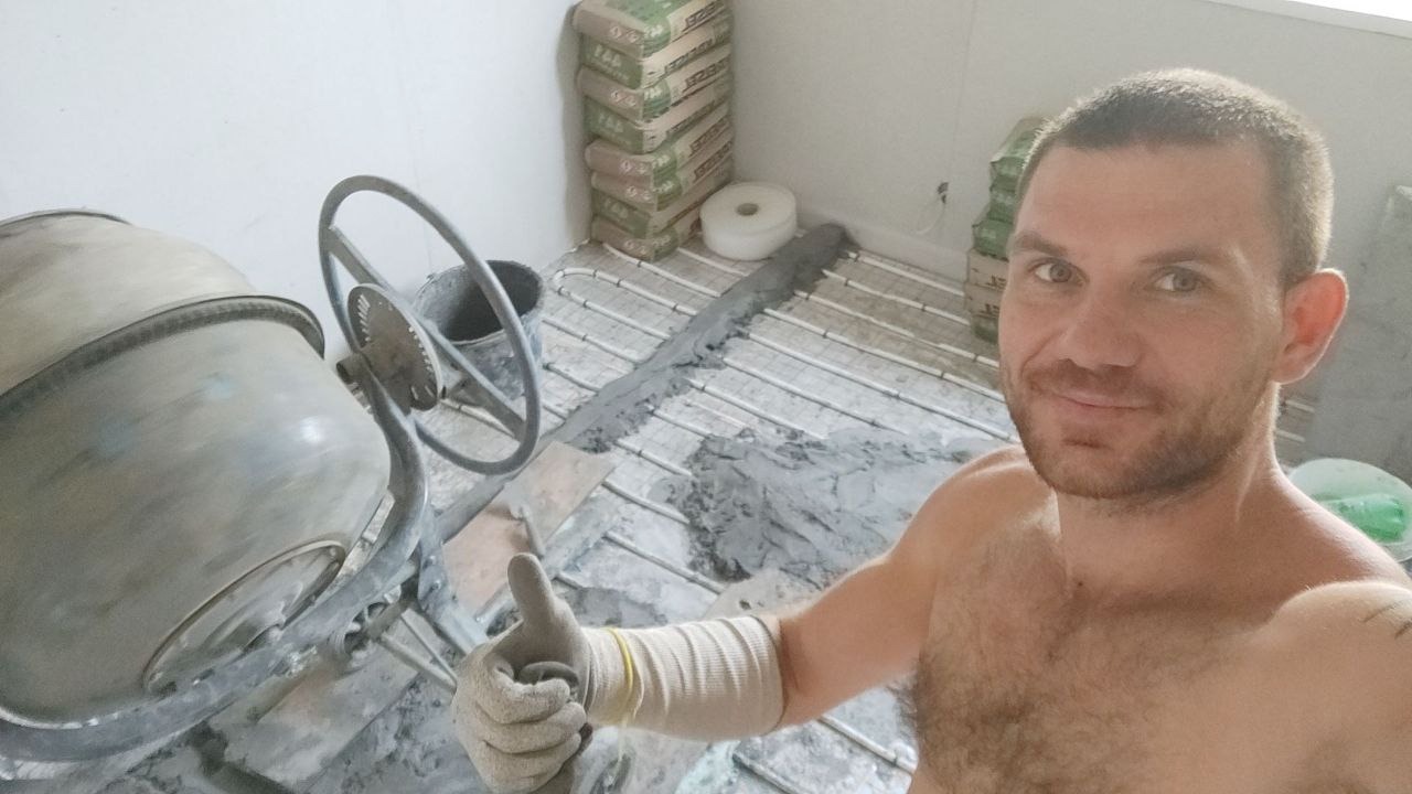 Mężczyzna z uniesionym kciukiem na tle betoniarki, worków z zaprawą i instalacji ogrzewania podłogowego. Widoczny etap prac przygotowawczych do wylewki.