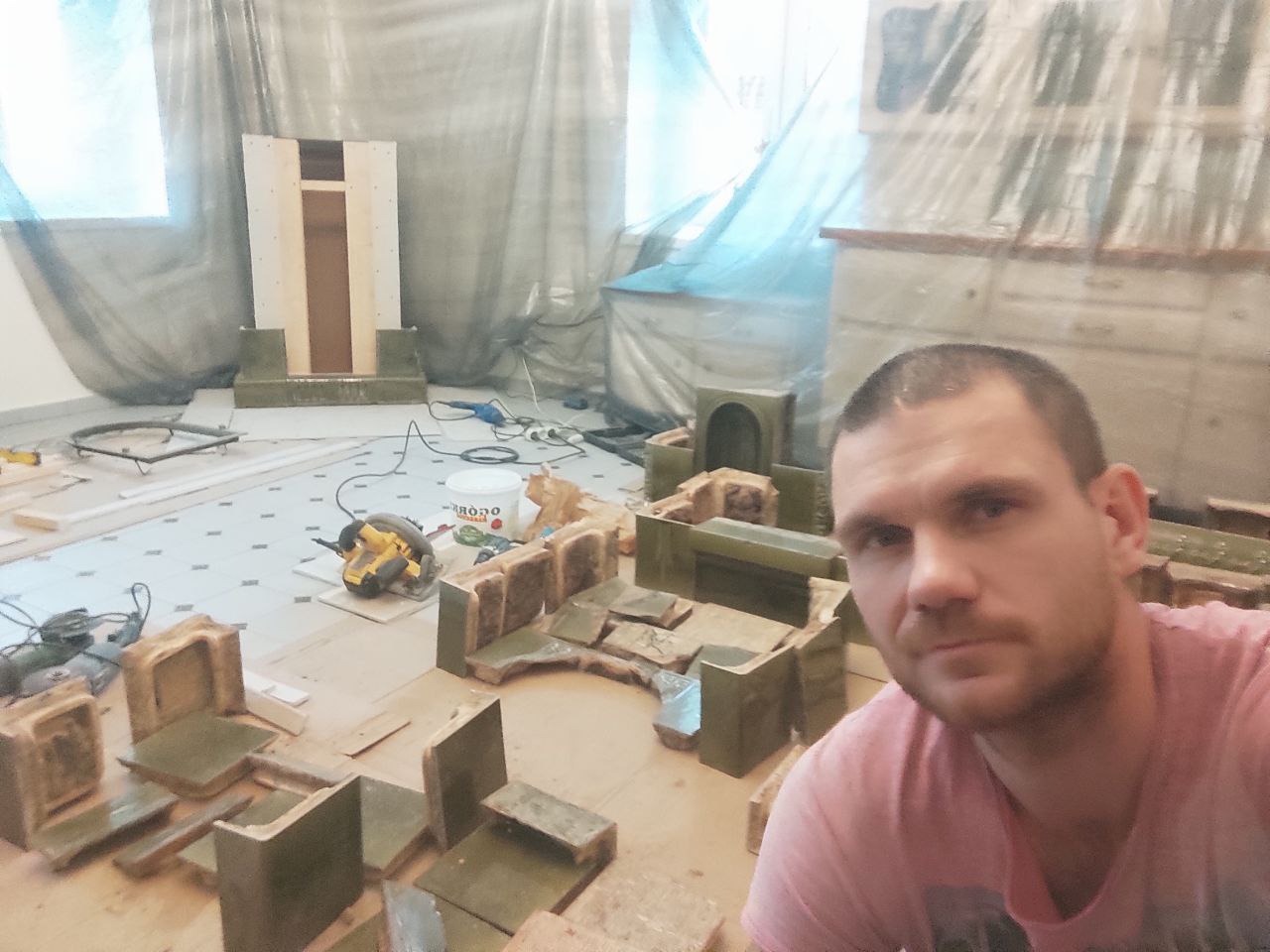 Montaż dekoracyjnego kominka z zielonych kafli w trakcie prac wykończeniowych. Widoczny wykonawca i narzędzia. Całość osłonięta folią malarską.