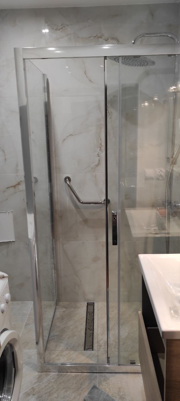 Nowoczesna kabina prysznicowa z chromowanymi detalami i odpływem liniowym, wykończona płytkami imitującymi marmur. Widoczna armatura i umywalka.