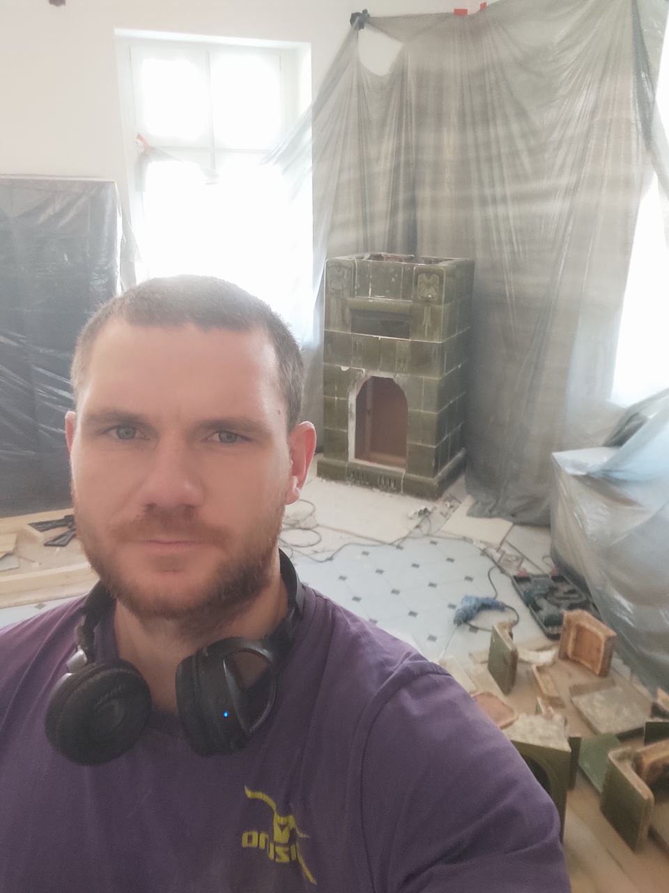 Mężczyzna z słuchawkami na szyi pozuje na tle remontowanego pomieszczenia z rozłożonymi kaflami i piecem kaflowym w trakcie budowy.