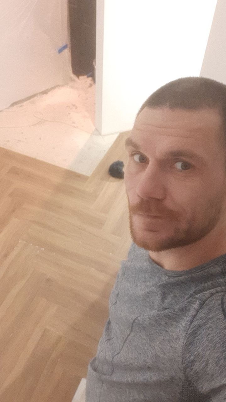 Parkiet ułożony w jodełkę, widok z góry z perspektywy osoby stojącej w pomieszczeniu w trakcie wykańczania. W tle fragment ściany i otwór drzwiowy.