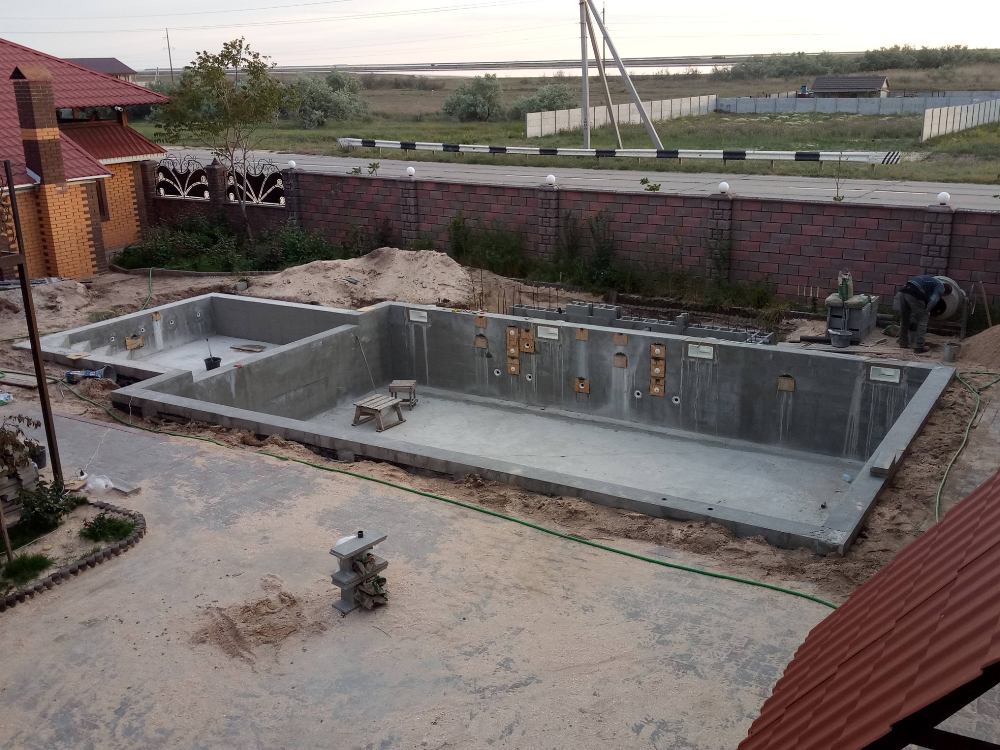 Budowa betonowego basenu na posesji: widok z góry na częściowo wypełniony wykop, z widocznymi otworami na dysze i pracującym mężczyzną przy betoniarce w tle.