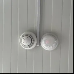 Biały czujnik dymu i czujnik ruchu zamontowane na białym, panelowym suficie. Widoczny fragment białej listwy montażowej.