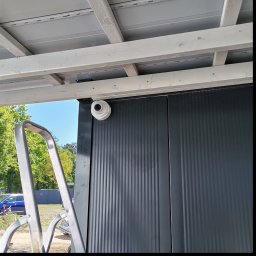 Pod Okiem - Kamera monitoringu kopułkowa, zamontowana na szarej, pionowo żebrowanej ścianie pod zadaszeniem. Obok drabina, w tle zieleń drzew i błękit nieba.