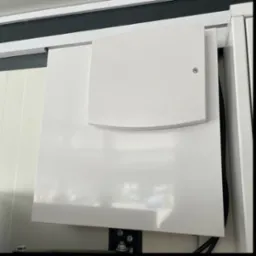 Zamknięta, biała skrzynka elektryczna z widocznymi przewodami, zamontowana na ścianie wewnątrz pomieszczenia. Widoczny fragment metalowej konstrukcji i śruby mocujące.