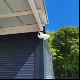 Zewnętrzna kamera monitoringu kopułkowa, zamontowana na rogu budynku z żaluzjami, na tle błękitnego nieba i zieleni drzew. Widok z dołu.