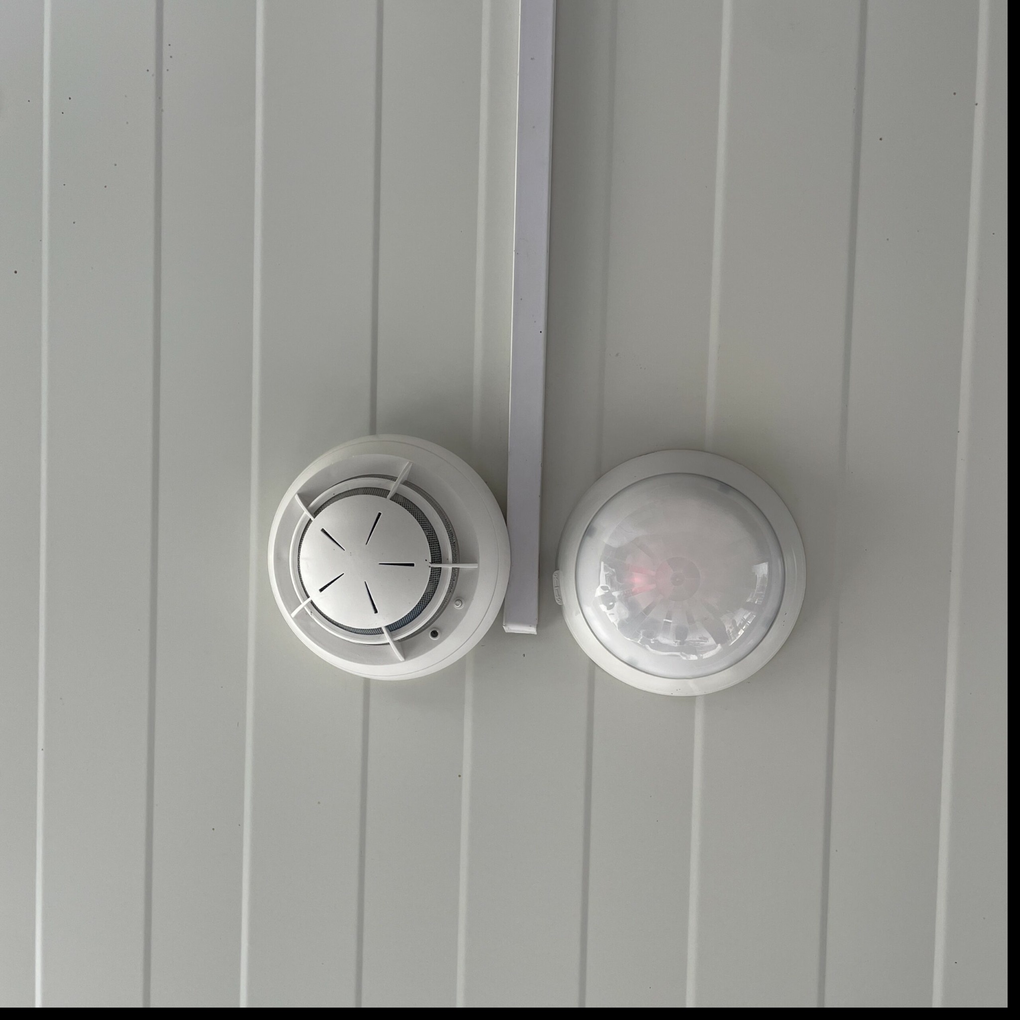 Biały czujnik dymu i czujnik ruchu zamontowane na białym, panelowym suficie. Widoczny fragment białej listwy montażowej.