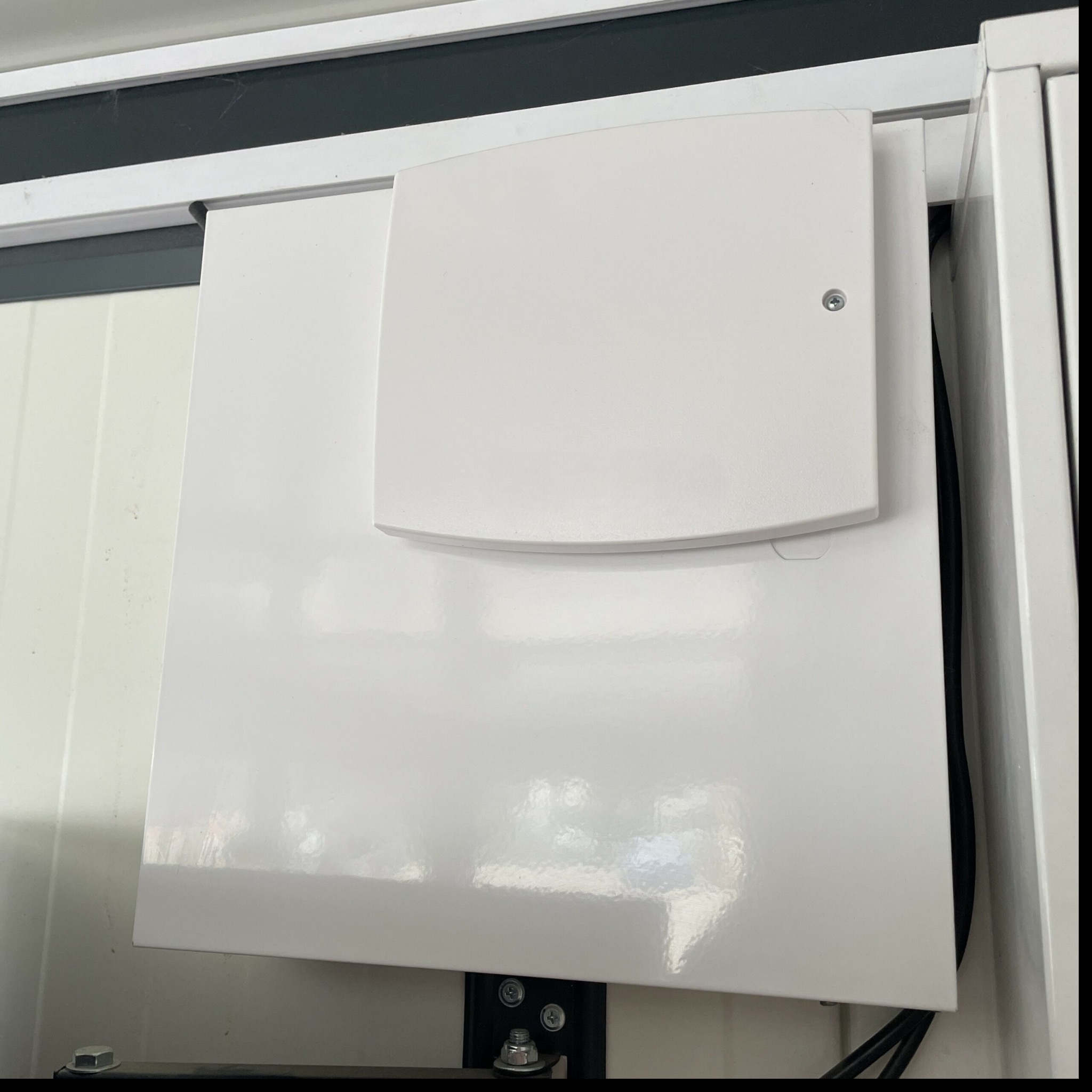 Zamknięta, biała skrzynka elektryczna z widocznymi przewodami, zamontowana na ścianie wewnątrz pomieszczenia. Widoczny fragment metalowej konstrukcji i śruby mocujące.