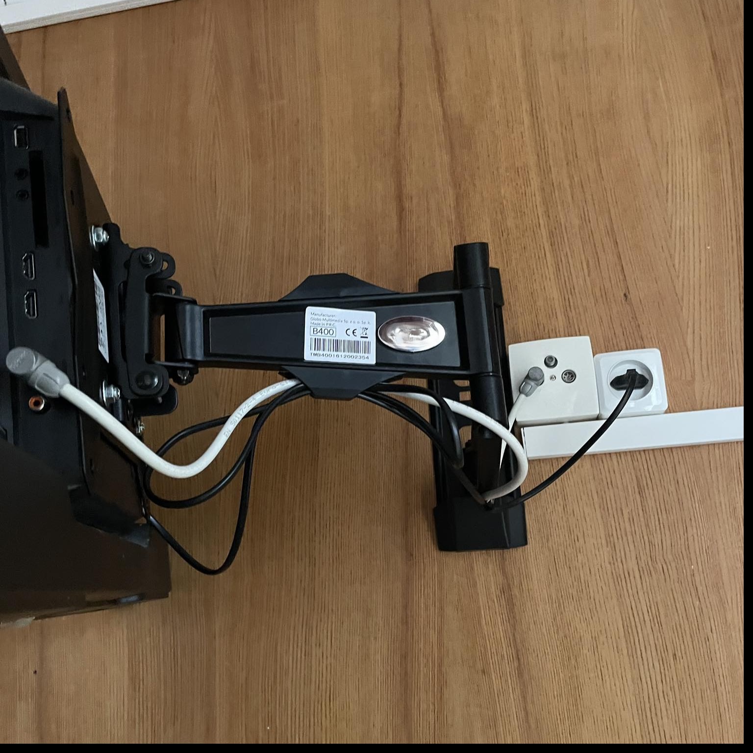 Zamontowany uchwyt do telewizora na drewnianej ścianie, widoczne gniazdka elektryczne i przewody. Czarny uchwyt z regulacją położenia ekranu.