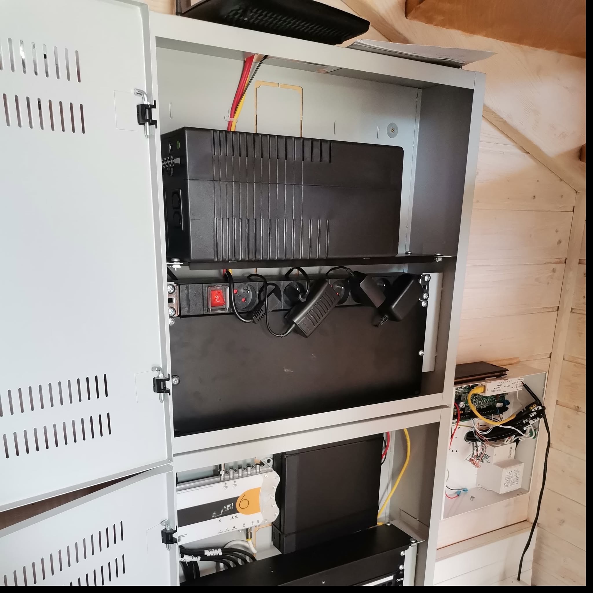 Zamknięta szafa rack z zainstalowanym sprzętem sieciowym, zasilaczem awaryjnym UPS i listwą zasilającą. Widoczne przewody i kable. Wnętrze wykończone boazerią.