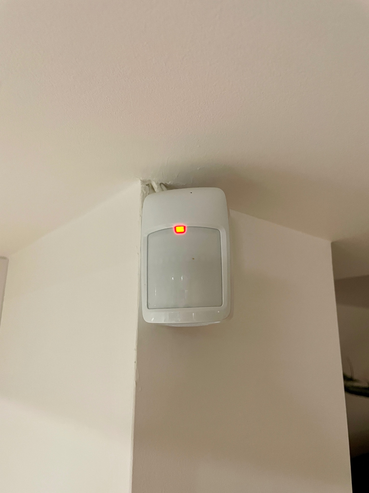 Zamontowany na rogu ściany czujnik alarmowy z czerwoną diodą, sygnalizujący gotowość systemu. Widoczne niedoskonałości wykończenia wokół urządzenia.