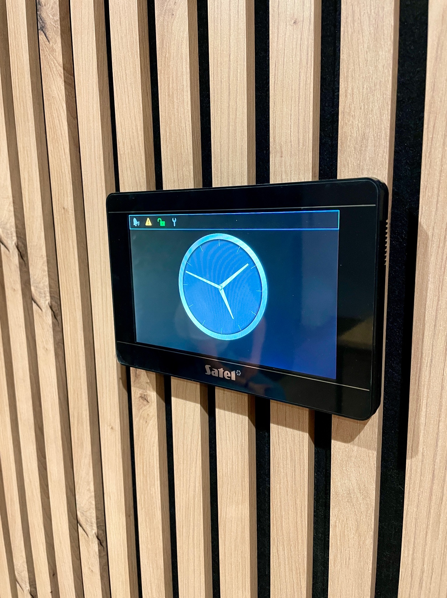 Panel sterowania systemem alarmowym Safel na drewnianej ścianie z pionowymi listwami. Widoczne ikony alarmu, klucza i narzędzi.