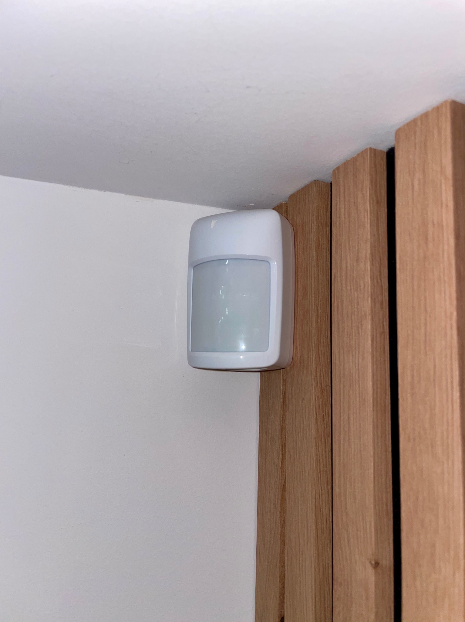 Zamontowany czujnik ruchu alarmowego, biały, prostokątny, na styku białej ściany i drewnianej framugi. Widok z dołu, blisko sufitu.