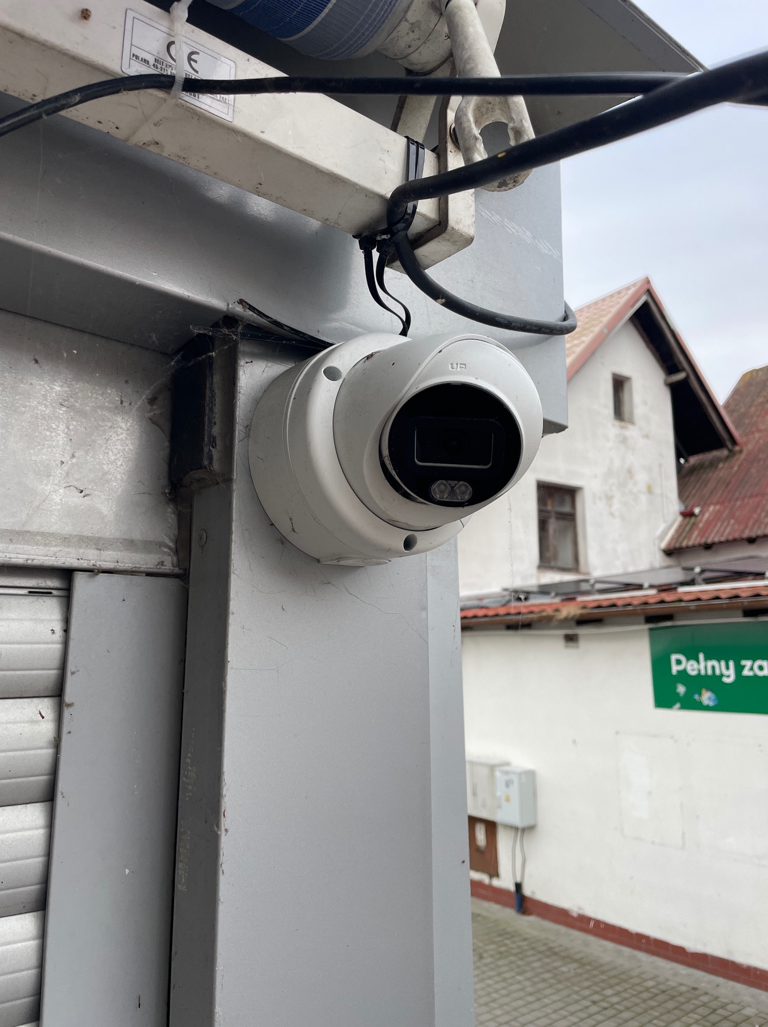 Kamera monitoringu kopułkowa, zamontowana na szarej elewacji budynku z widocznymi przewodami. W tle budynek mieszkalny i szyld reklamowy.