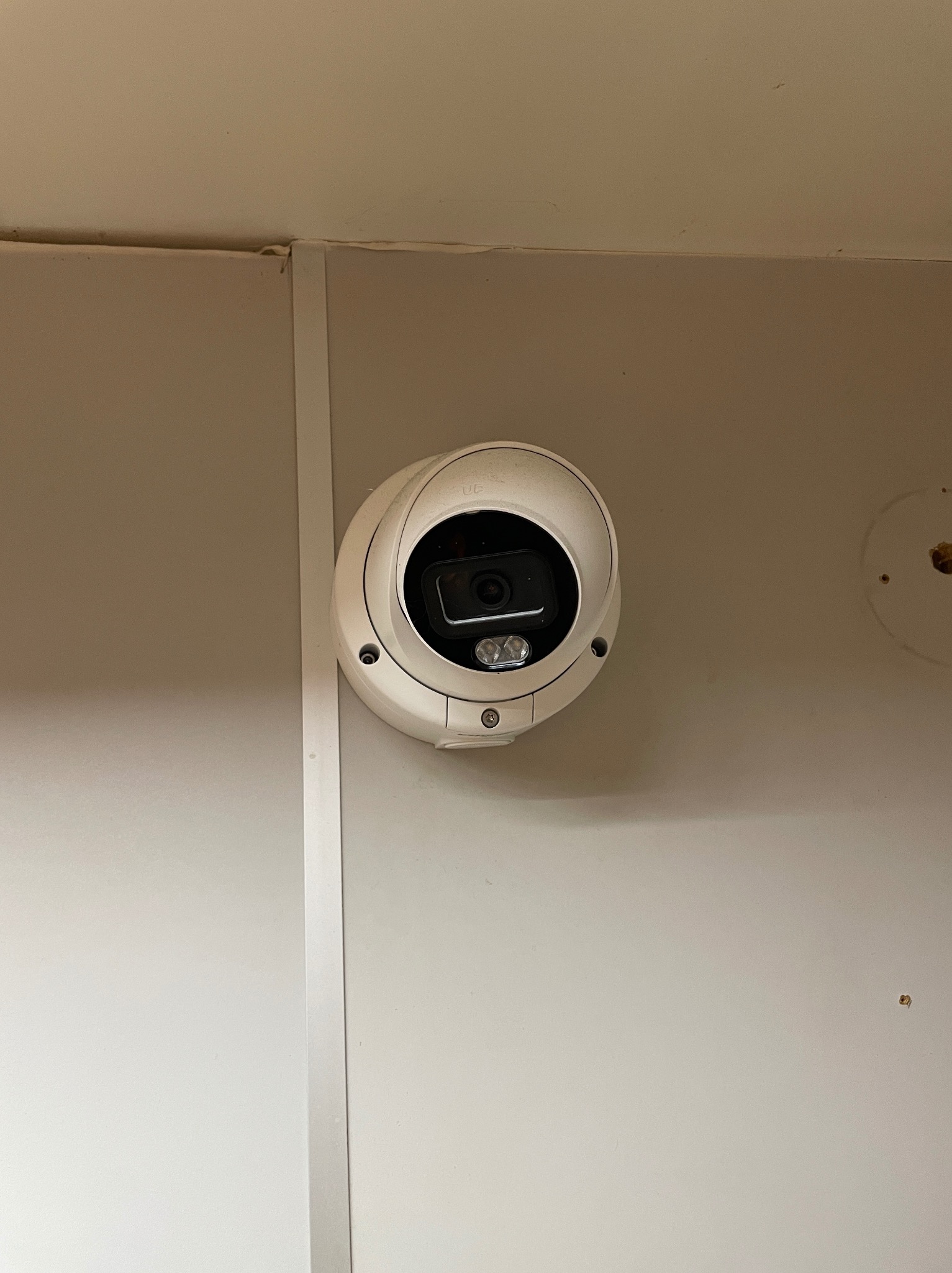 Kamera kopułkowa monitoringu, zamontowana na jasnej ścianie. Widoczne ślady po poprzednim montażu obok kamery. Ujęcie z bliska, centralne.