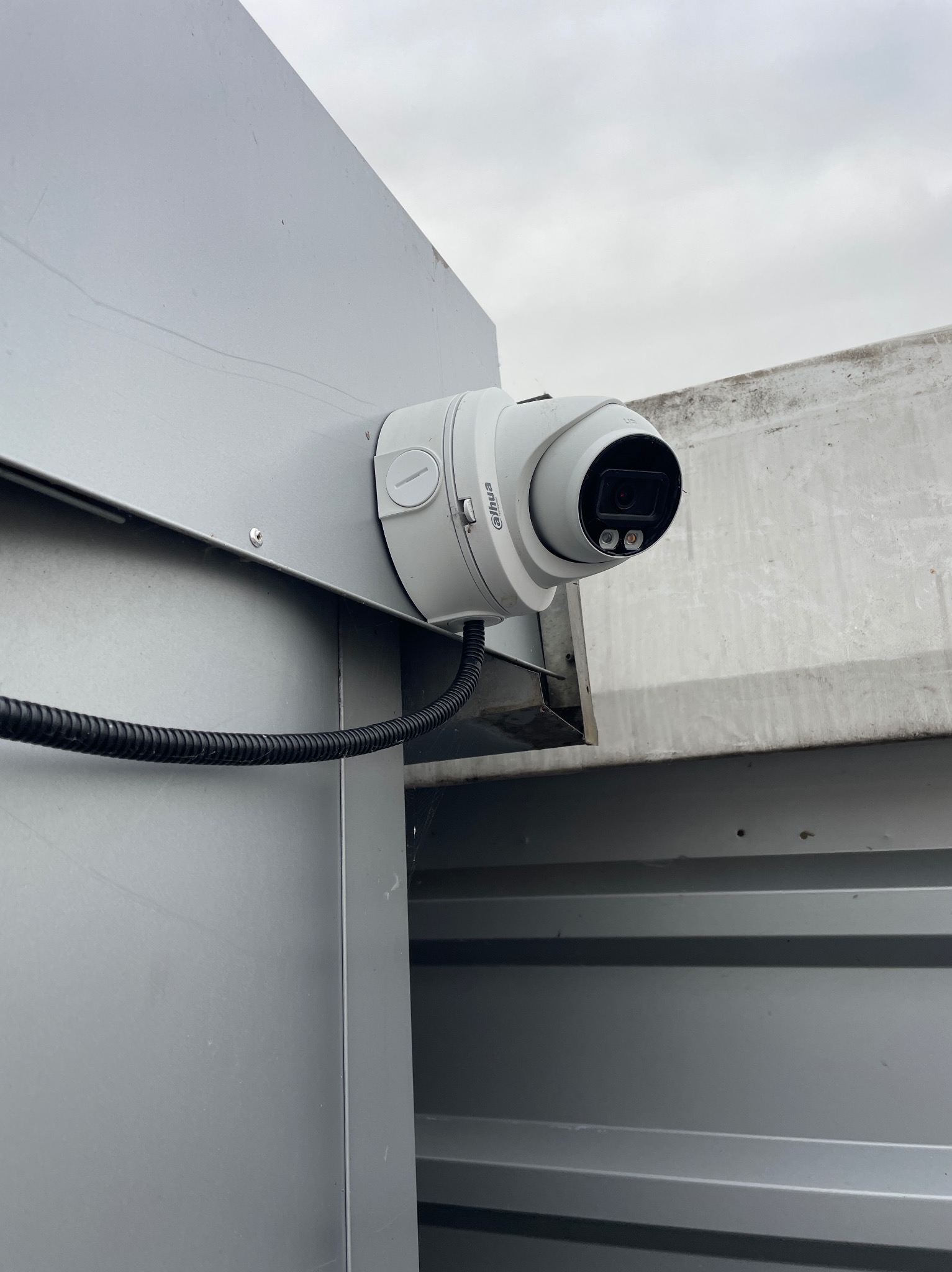 Kamera monitoringu Dahua zamontowana na szarej, metalowej elewacji budynku, widoczna z dołu pod kątem. Kabel w peszlu. Pochmurne niebo w tle.