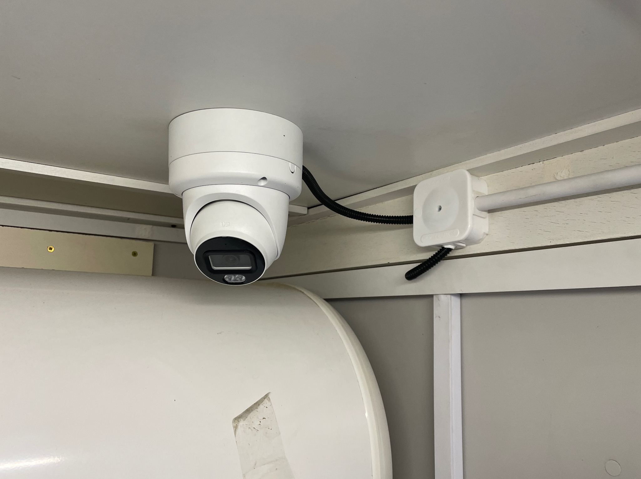 Kamera monitoringu kopułkowa, biała, zamontowana pod sufitem, z widocznym kablem i puszką instalacyjną. Ujęcie z bliska, ukazujące szczegóły montażu.