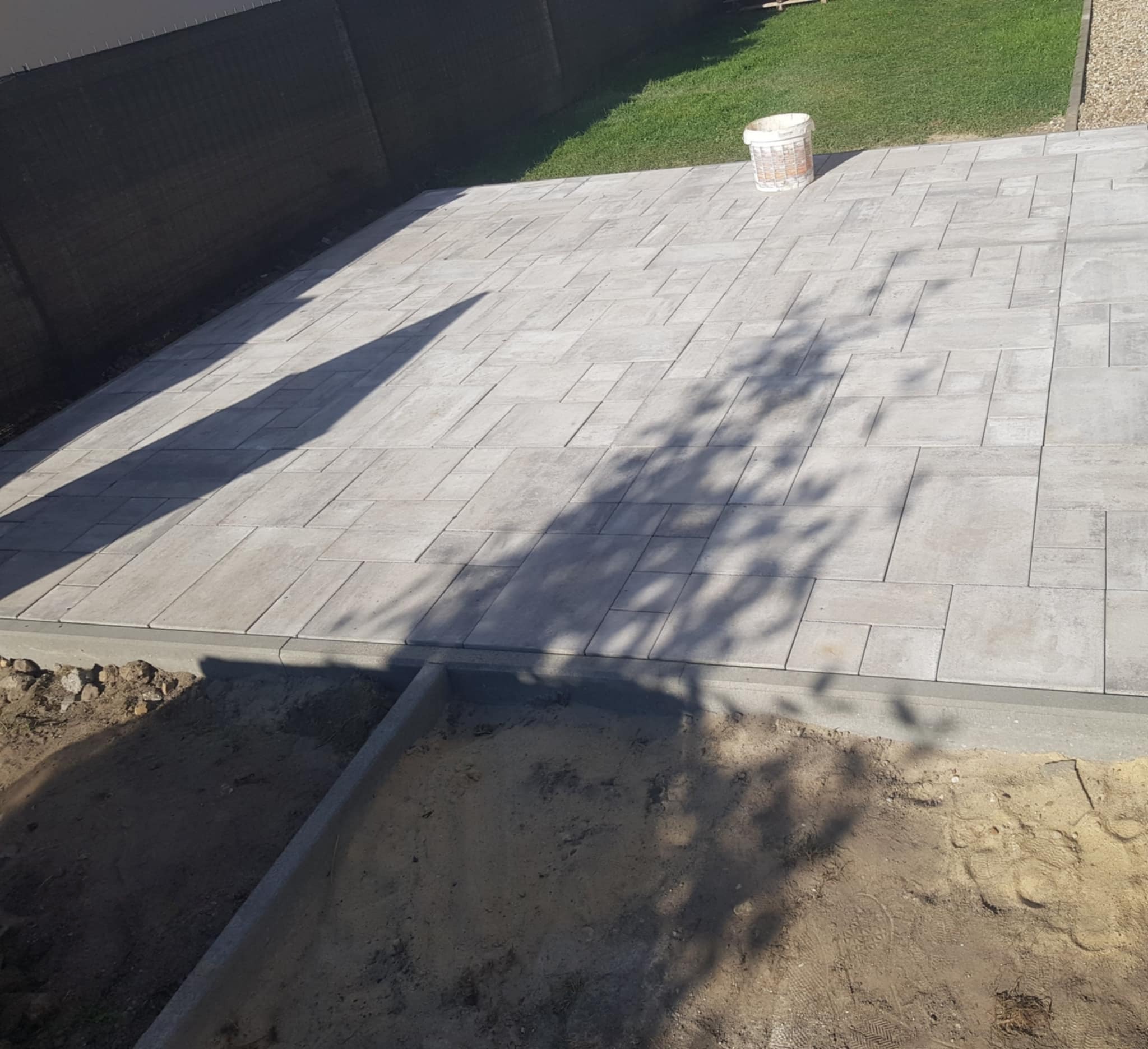 Szara kostka brukowa ułożona na podwórku, widok z góry. W tle siatka ogrodzeniowa i trawa, po prawej wiadro. Cień drzewa na kostce.