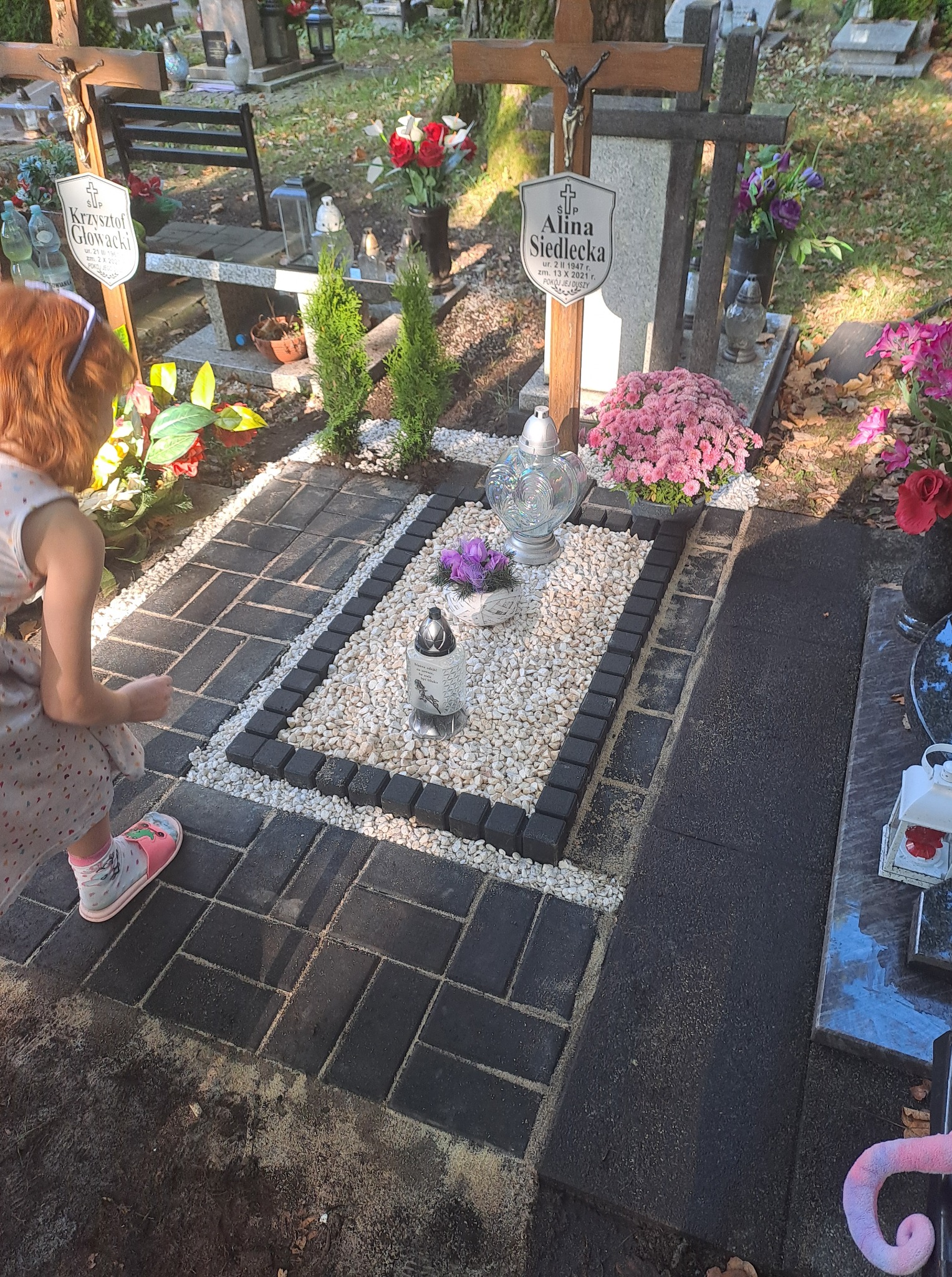 Wykończony nagrobek z czarnej kostki brukowej, obramowany białymi kamykami i czarnymi kostkami. Dziewczynka przygląda się nagrobkowi.