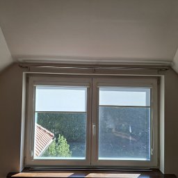 Design & Programming Chyliński Mariusz - Okno dachowe z roletą w pokoju na poddaszu, widok na zieleń i dach z czerwonej dachówki. Metalowy karnisz nad oknem. Jasne ściany i sufit.